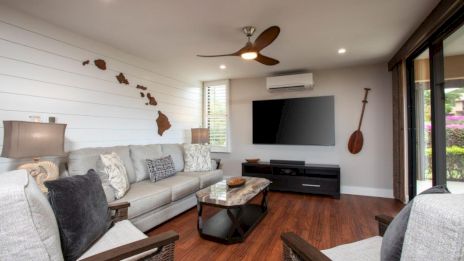 A cozy living room with a gray sofa, wooden floor, TV, wall decor, and a ceiling fan, creating a modern and welcoming atmosphere.