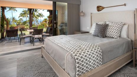 A modern bedroom with a neutral palette overlooks a tropical patio through large glass doors, featuring patterned bedding and an oar wall decor.