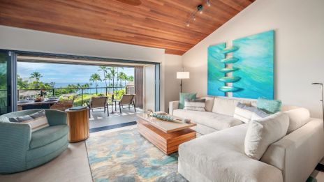 A modern living room with a large sofa, armchair, abstract art, and a view of an outdoor patio with palm trees and ocean scenery.