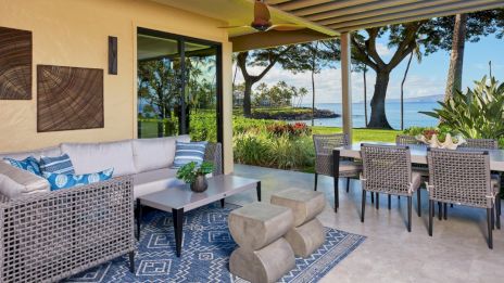 A patio with modern outdoor furniture, plants, and a scenic ocean view, featuring a blue patterned rug and decorative wall art.