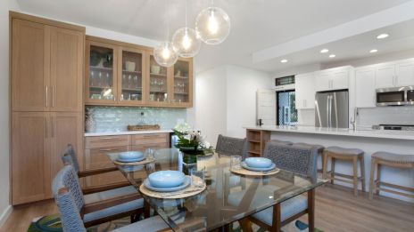 A modern dining area with a glass table, blue plates, and wicker chairs; a kitchen with wooden cabinets and pendant lights is visible.