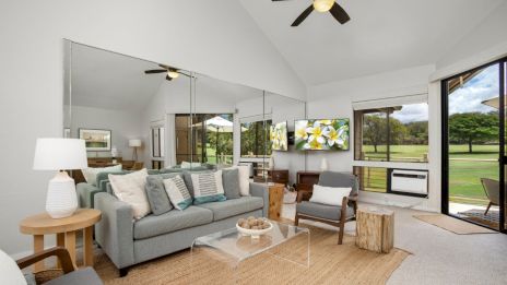 A modern living room with a sofa, armchairs, mirrors, TV, ceiling fan, and large windows overlooking a grassy outdoor area.