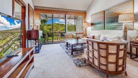 A cozy living room features a TV, sofa, and coffee table, with large windows offering a scenic view and outdoor seating area.