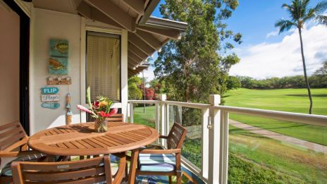 A wooden balcony set overlooks a lush golf course. There's a vase with flowers, a flip-flop themed sign, trees, and a palm tree.