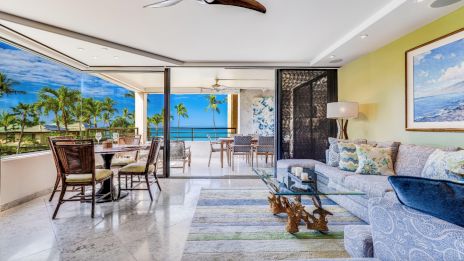 A luxurious living room with a glass wall opens to an ocean-view balcony. It features elegant furnishings and tropical decor.