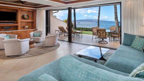 A spacious living room with turquoise seating opens to a patio with chairs, overlooking the ocean and palm trees.