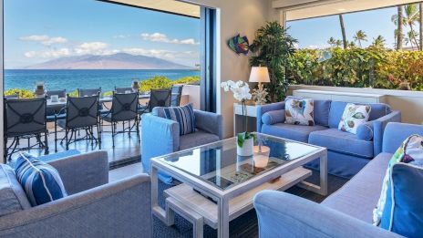 A bright living room with blue sofas and a glass table, offering a stunning ocean view and outdoor seating.