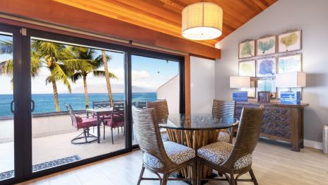Room with a round dining table, ocean view through large windows, palm trees outside, wooden ceiling, and wall art above a dresser.