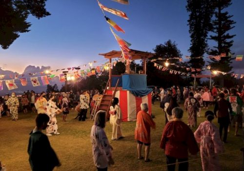 A vibrant festival with people in traditional attire, a decorated platform, and festive lights, set against a dusk sky.