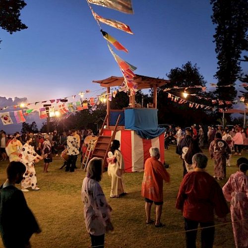 A vibrant festival with people in traditional attire, a decorated platform, and festive lights, set against a dusk sky.
