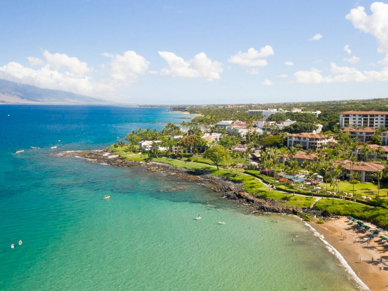 A scenic coastal view featuring a sandy beach, clear turquoise waters, lush greenery, and buildings under a partly cloudy sky with distant mountains.