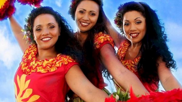 Three people in traditional Hawaiian attire, smiling and holding red-and-yellow pom-poms against a blue sky backdrop.