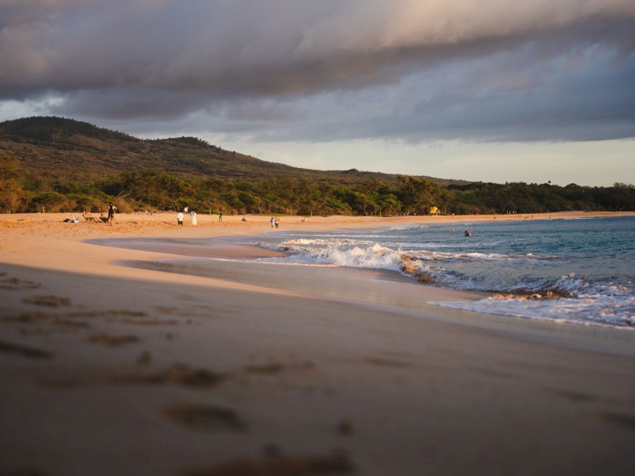 A serene beach scene with gentle waves, scattered footprints on the sand, and a backdrop of lush hills under a cloudy sky.