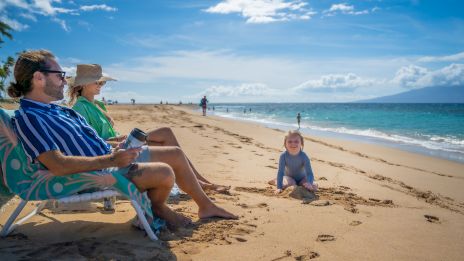 A couple relaxes on a beach while a child plays in the sand near the ocean under a clear blue sky.