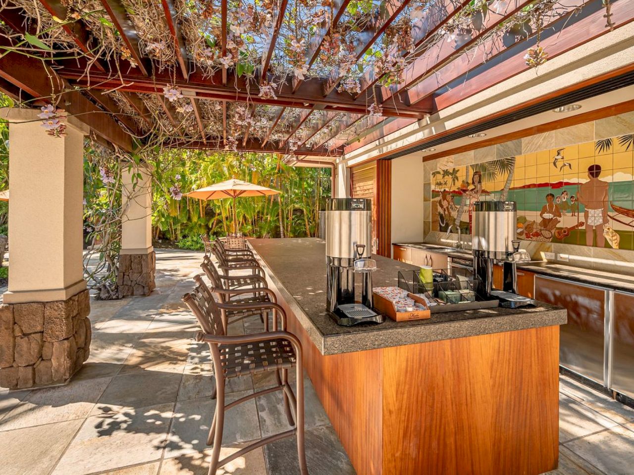 Outdoor bar with wooden counter and stools under a pergola. A mural decorates the wall. Tables set in a sunny, tropical setting.