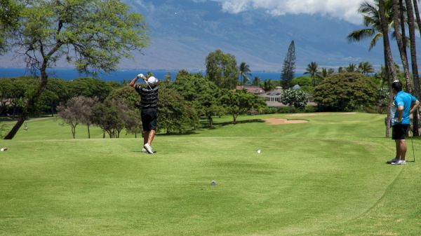 Two people are on a golf course; one is swinging a club, while the other watches. Lush greenery and mountains are in the background.