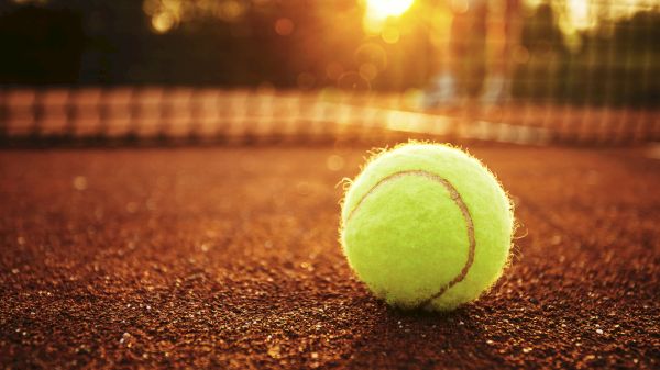 A tennis ball lies on a clay court, with the net and a sunset in the background, creating a warm and serene scene.