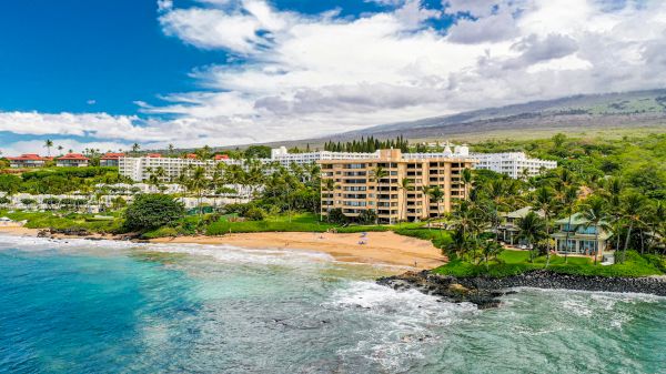 A beachfront resort with lush greenery, a sandy beach, and mountains in the background under a partly cloudy sky.