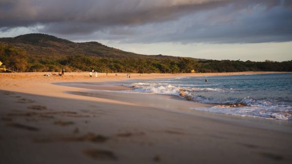 A sandy beach with gentle waves, surrounded by hills under an overcast sky.
