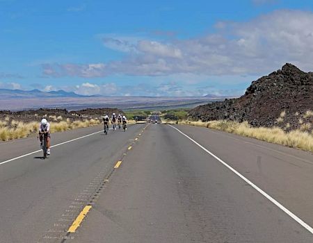 Cyclists ride along a long, straight desert highway flanked by dry grasses and rocky outcrops under a blue sky with scattered clouds.