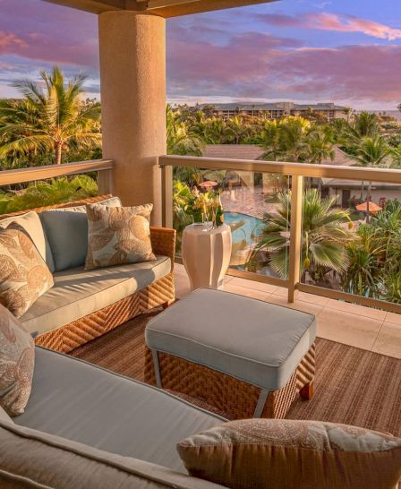 A cozy patio with cushioned seating overlooks a lush, tropical landscape with palm trees and a pool under a vibrant, colorful sky.