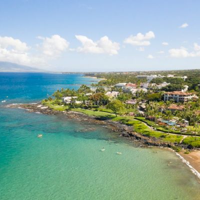 Aerial view of a coastal area with clear blue water, sandy beaches, and lush greenery near buildings and mountains under a partly cloudy sky.