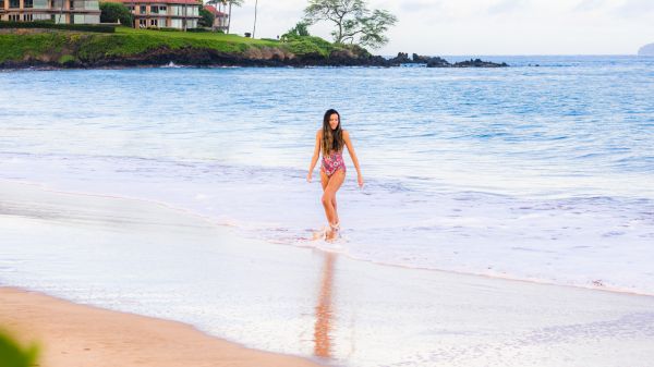A person walking along a beach with gentle waves, near oceanfront homes surrounded by greenery and palm trees.