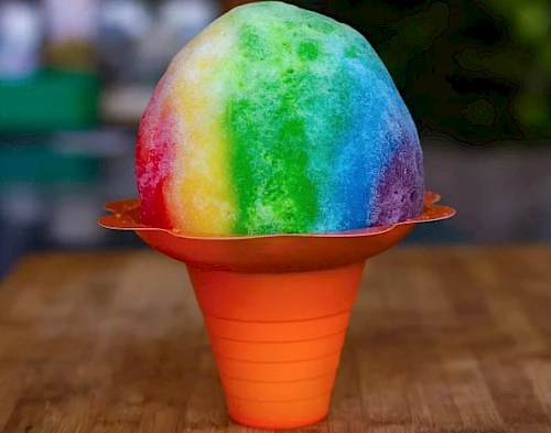 A vibrant rainbow-colored snow cone in an orange holder on a wooden surface.