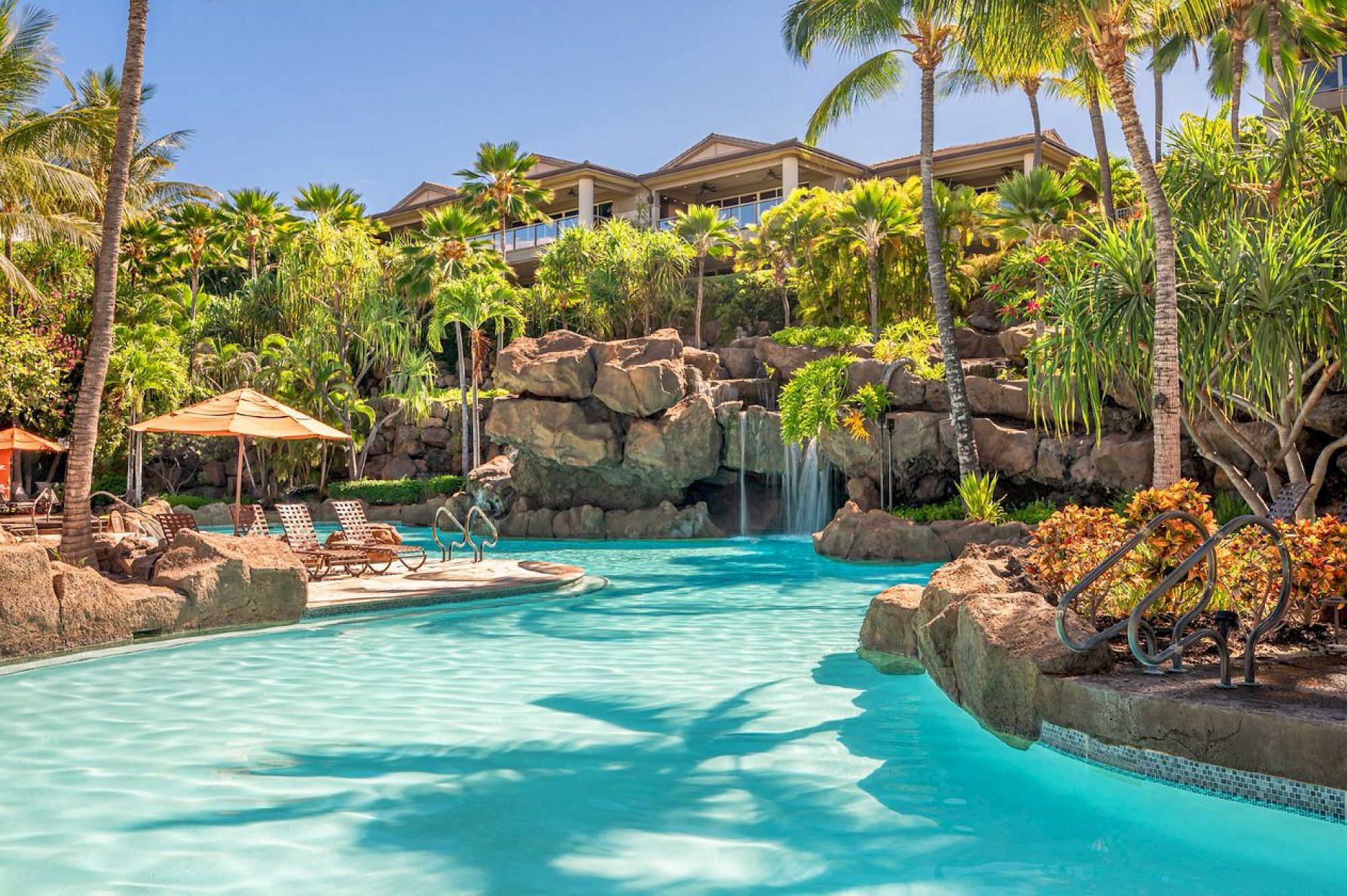 A tropical resort pool with lounge chairs, umbrellas, and lush greenery, featuring a waterfall and a rocky backdrop under a clear sky.