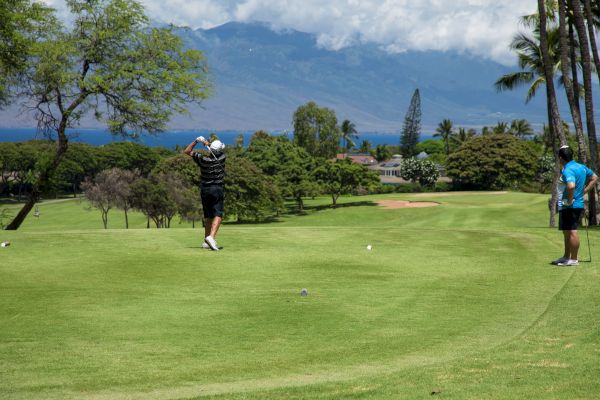Two people on a golf course, with one swinging a club and the other watching, surrounded by trees and mountains in the background.