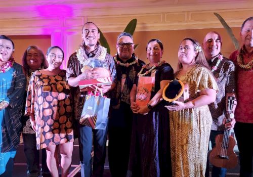 A group of people dressed in colorful attire, some holding gifts, standing together indoors with decorative lighting.