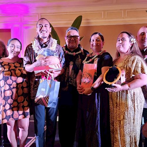 A group of people dressed in colorful attire, some holding gifts, standing together indoors with decorative lighting.