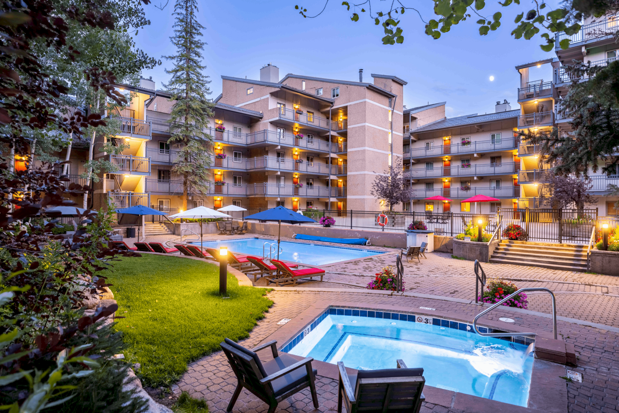 A cozy outdoor area with a pool, hot tub, lounge chairs, and illuminated multi-story buildings under a twilight sky.