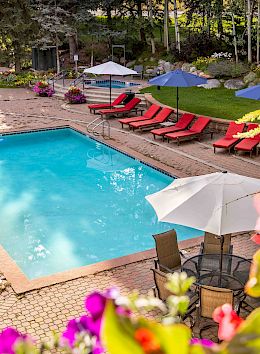 A swimming pool surrounded by red lounge chairs, blue umbrellas, and vibrant flowers in a peaceful outdoor setting.