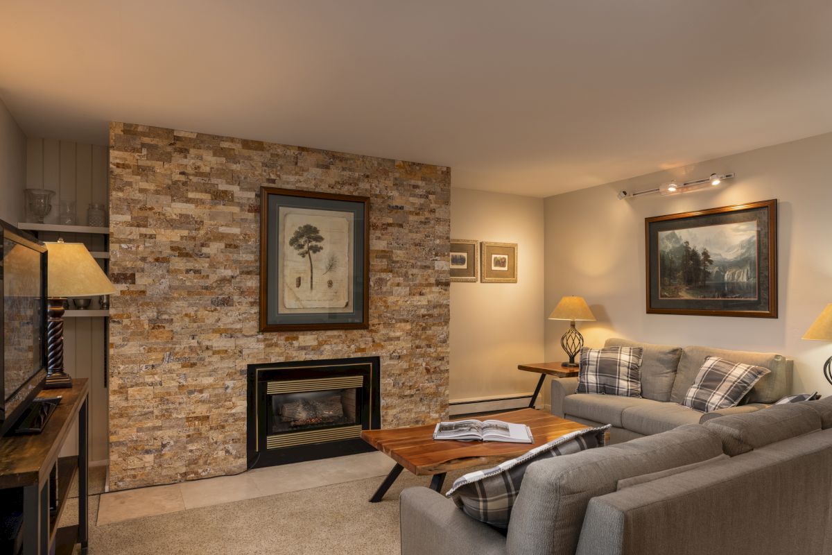 The image shows a cozy living room with a stone feature wall, art pieces, comfortable sofas, a coffee table, and warm lighting.