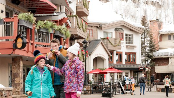 A winter scene with three people in colorful jackets enjoying treats in a snowy village, with charming buildings in the background.