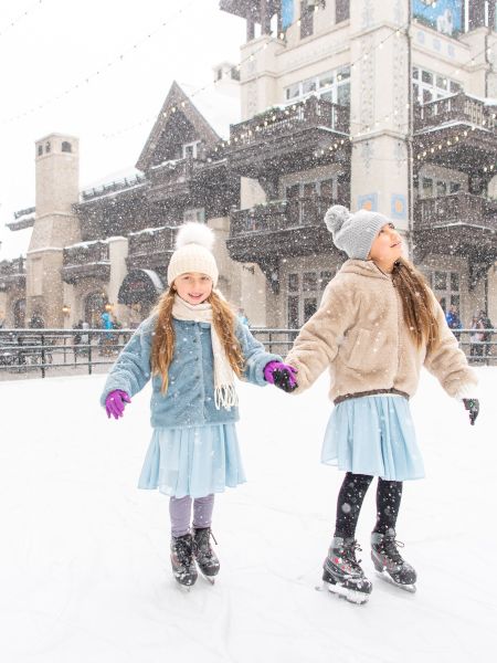 Two girls in winter attire are ice skating outdoors, holding hands, with snow falling around them in a charming town setting.