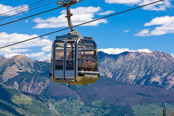 The image features a gondola cable car traveling over lush mountains under a bright blue sky with clouds.