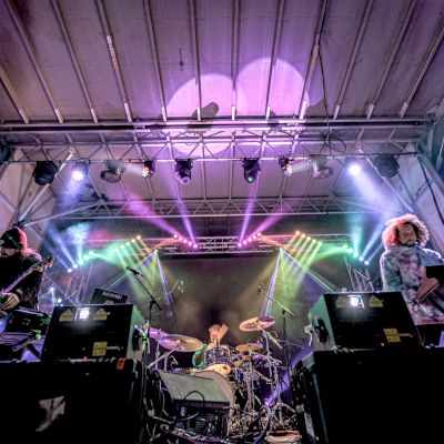 A band performs live on stage, with colorful lights and musical equipment surrounding them, creating an energetic concert atmosphere.