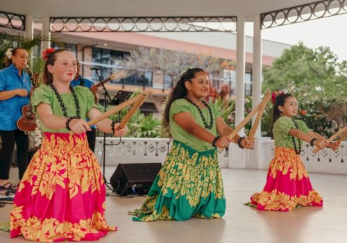 Three young dancers in colorful skirts perform with sticks, accompanied by a musician on stage in an outdoor setting.