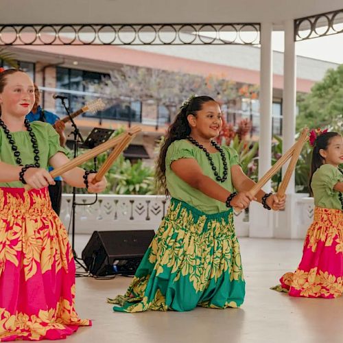 Three young dancers in colorful skirts perform with sticks, accompanied by a musician on stage in an outdoor setting.