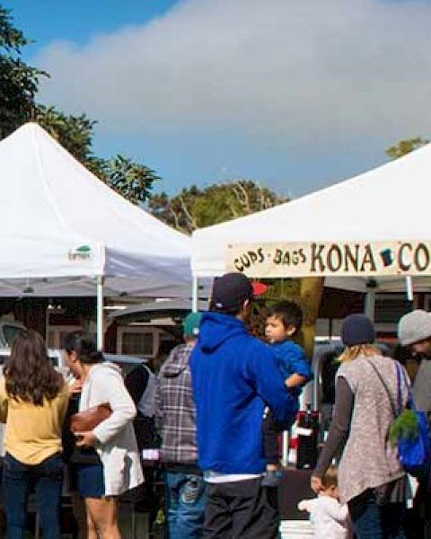 People are gathered around tents at an outdoor market or festival, with one tent labeled for Kona coffee.