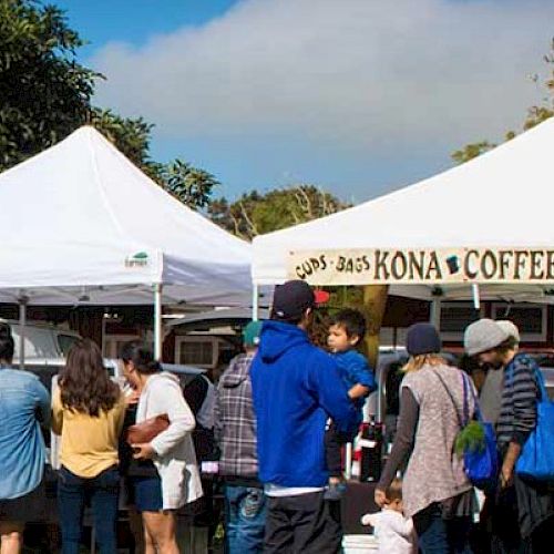 People are gathered around tents at an outdoor market or festival, with one tent labeled for Kona coffee.