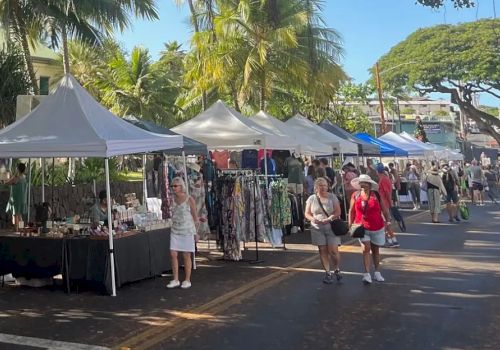 A street market with people browsing stalls selling clothes and various items, under tents with palm trees lining the road.