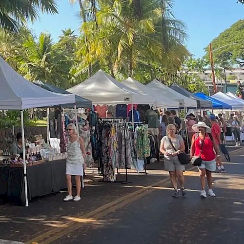 A street market with people browsing stalls selling clothes and various items, under tents with palm trees lining the road.