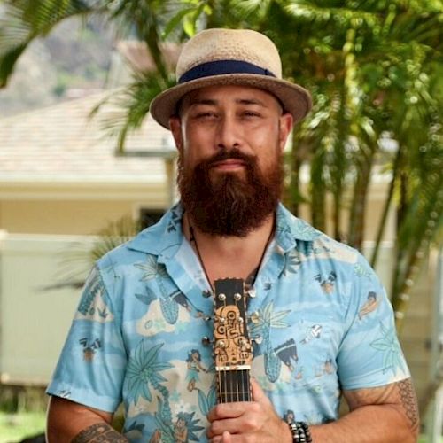 A person with a beard, wearing a hat and a tropical shirt, stands with a string instrument outdoors near palm trees.