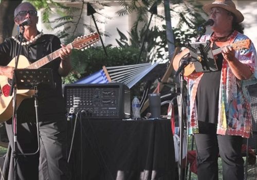 Two musicians play acoustic guitars on a sunny outdoor stage, singing into mics with a rolling booth of gear beside them.