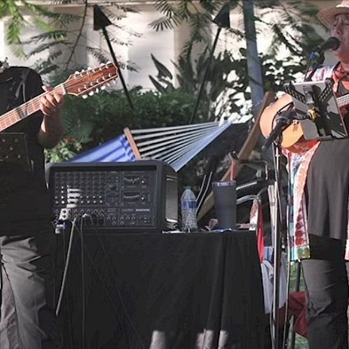 Two musicians play acoustic guitars on a sunny outdoor stage, singing into mics with a rolling booth of gear beside them.