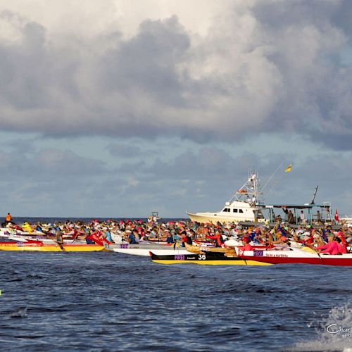 The image depicts a group of colorful canoes racing on the ocean with several boats in the background under a cloudy sky.