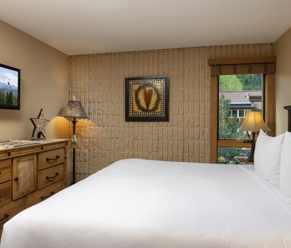 A cozy bedroom features a large bed, wooden dresser, two lamps, and art pieces on the wall, with a window offering a scenic view outside.